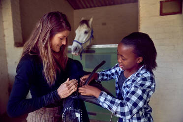 Frau hilft Mädchen bei der Vorbereitung des Sattels im Pferdestall - CAIF24291
