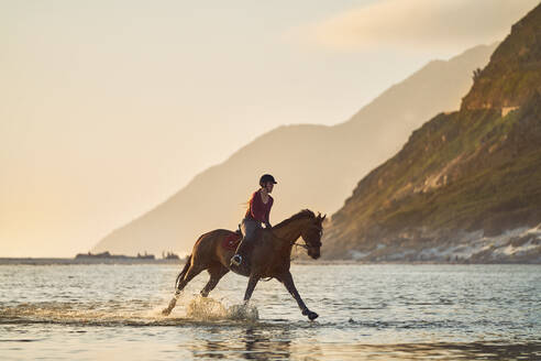 Junge Frau im Galopp auf einem Pferd in der ruhigen Meeresbrandung - CAIF24319