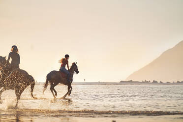 Junge Frauen reiten in der Meeresbrandung bei Sonnenuntergang - CAIF24336