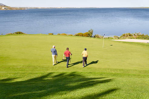Männliche Golfer gehen auf dem sonnigen Putting Green am See auf den Pin zu - CAIF24498