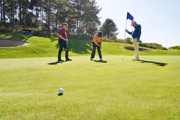 Männliche Golffreunde beim Putten zu Hause auf dem sonnigen Golfplatz - CAIF24510