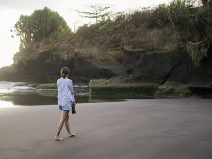 Junge Frau geht an einem felsigen Strand spazieren und schaut - CAVF74988