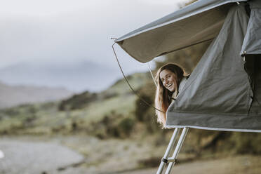 Eine junge lächelnde Frau streckt ihren Kopf aus einem Zelt in Neuseeland. - CAVF74992