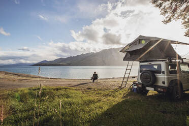 Eine Frau sitzt an einem See neben einem Geländewagen mit Zelt in Neuseeland - CAVF74993