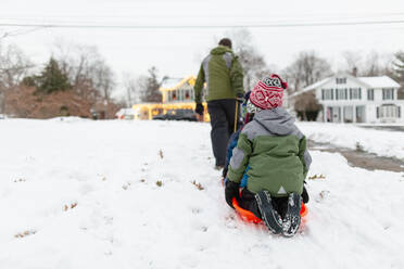 Rückansicht eines Vaters, der seine Kinder auf einem orangefarbenen Schlitten durch den Schnee zieht - CAVF75006