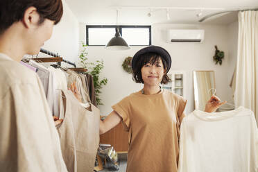 Two Japanese women standing in a small fashion boutique, looking at tops. - MINF13722