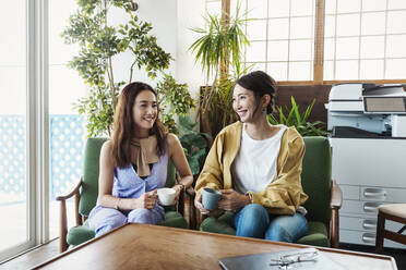 Two female Japanese professionals sitting in a co-working space, smiling at each other. - MINF13745