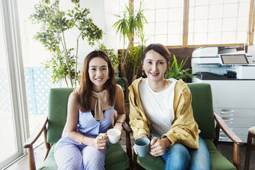 Two female Japanese professionals sitting in a co-working space, smiling at camera. - MINF13746
