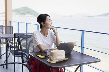 Female Japanese professional sitting on balcony of a co-working space, using laptop computer. - MINF13770