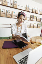 Female Japanese professional sitting at table in a co-working space, using digital tablet and laptop, holding book. - MINF13773