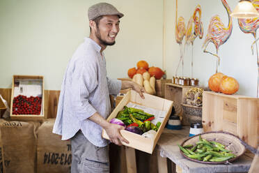 Smiling Japanese man wearing cap standing in farm shop, holding crate with fresh vegetables. - MINF13804