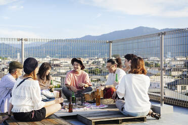 Eine Gruppe junger Japanerinnen und Japaner sitzt auf einer Dachterrasse in einer städtischen Umgebung und trinkt Bier. - MINF13808