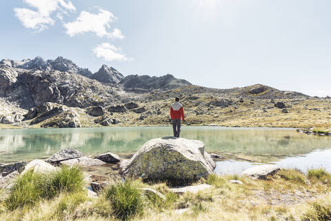 Rückansicht eines jungen Mannes auf einem Felsen mit Blick auf einen klaren Bergsee, lizenzfreies Stockfoto