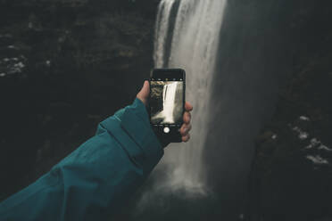 Junger Mann fotografiert einen Wasserfall mit seinem Smartphone - CAVF75060