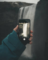 Abgeschnittene Hand beim Fotografieren eines Wasserfalls in Island mit einem Smartphone - CAVF75061