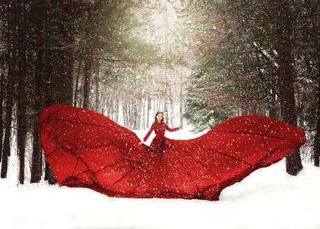 Young redhead girl in flowing red dress in forest with snowfall. - CAVF75352