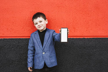 Portrait of content little boy wearing suit coat showing smartphone - LJF01314