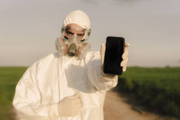 Mann mit Schutzanzug und Maske auf dem Land, der ein Mobiltelefon hält - ERRF02628