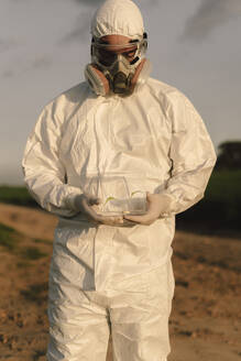 Mann mit Schutzanzug und Maske auf dem Lande, der einen Setzling in einem Glaskasten hält - ERRF02638