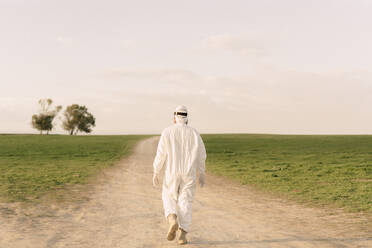 Rear view of man wearing protective suit walking on dirt track in the countryside - ERRF02639