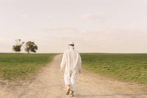 Rear view of man wearing protective suit walking on dirt track in the countryside - ERRF02639
