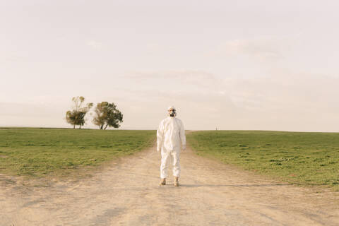 Mann mit Schutzanzug und Maske steht auf einem Feldweg in der Natur, lizenzfreies Stockfoto