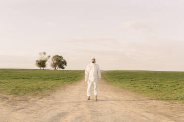 Man wearing protective suit and mask standing on dirt track in the countryside - ERRF02640