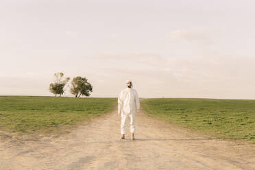 Mann mit Schutzanzug und Maske steht auf einem Feldweg in der Natur - ERRF02640