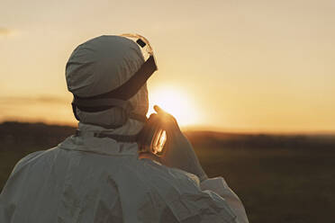 Rear viewof man wearing protective suit and mask in the countryside at sunset - ERRF02671