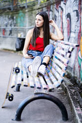 Young woman sitting on a bench and holding smartphone at skatepark - GIOF08029