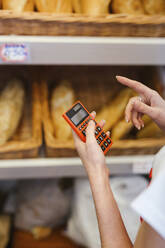 Junge Frau arbeitet in einer Bäckerei und benutzt einen Taschenrechner - LJF01343