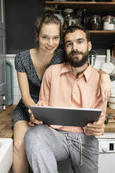 Young couple sitting on kitchen worktop, using digital tablet - PESF01809