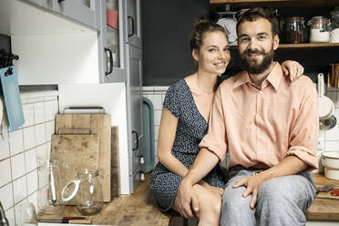 Junges Paar sitzt lächelnd auf der Küchenarbeitsplatte - PESF01811
