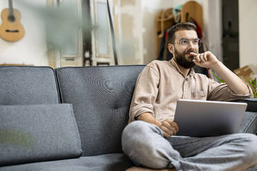 Young man sitting at home on couch, using digital tablet - PESF01817