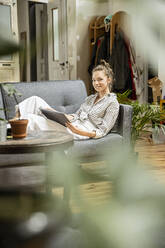 Young woman sitting at home on couch, using digital tablet - PESF01829