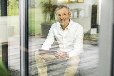 Smiling senior man with grey hair in modern design living room with tablet sitting on couch - SBOF02087