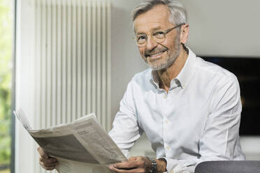 Porträt eines lächelnden älteren Mannes mit grauen Haaren in einem modern gestalteten Wohnzimmer, der Zeitung liest - SBOF02092