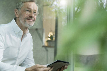 Smiling senior man with grey hair in modern design living room holding tablet - SBOF02099