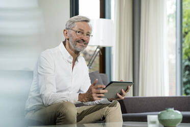 Smiling senior man with grey hair in modern design living room sitting on couch holding tablet - SBOF02101