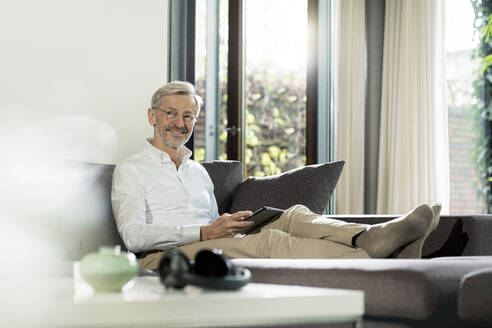 Lächelnder älterer Mann mit grauen Haaren in modernem Design Wohnzimmer sitzt auf Couch hält Tablet - SBOF02103
