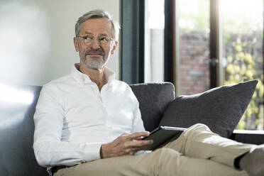 Älterer Mann mit grauen Haaren in modernem Design Wohnzimmer sitzt auf Couch hält Tablet - SBOF02104