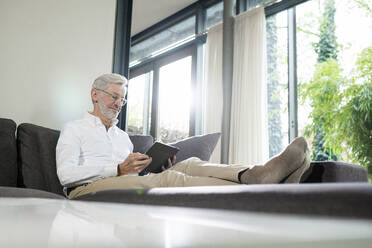 Älterer Mann mit grauen Haaren in modernem Design Wohnzimmer sitzen auf Couch mit Tablet - SBOF02106