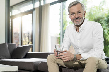 Porträt eines lächelnden älteren Mannes in einem modernen Design-Wohnzimmer, der auf einer Couch sitzt und ein Glas Wasser hält - SBOF02108