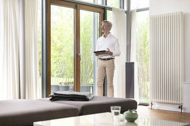 Senior man with grey hair in modern design living room standing at window holding tablet - SBOF02110