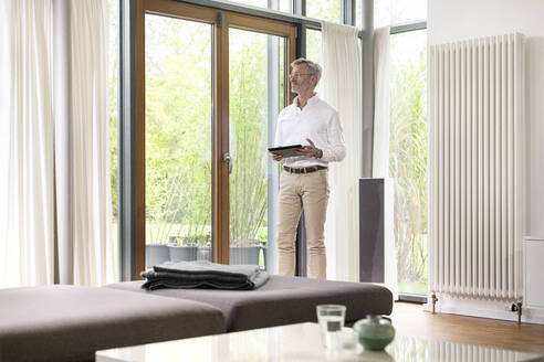 Älterer Mann mit grauen Haaren in modernem Design-Wohnzimmer am Fenster stehend mit Tablet - SBOF02110