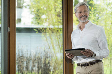 Lächelnder älterer Mann mit grauen Haaren in einem modern gestalteten Wohnzimmer, der am Fenster steht und ein Tablet hält - SBOF02113