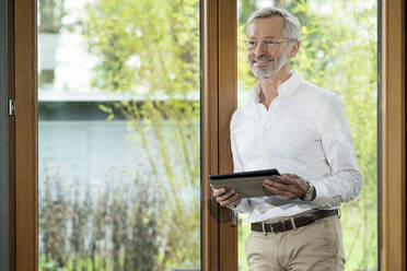 Lächelnder älterer Mann mit grauen Haaren in einem modern gestalteten Wohnzimmer, der am Fenster steht und ein Tablet hält - SBOF02114