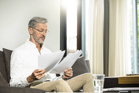 Senior Mann mit grauen Haaren in modernem Design Wohnzimmer sitzen auf Couch arbeiten auf Papiere im Home-Office, lizenzfreies Stockfoto