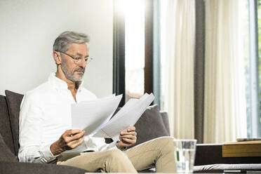 Senior Mann mit grauen Haaren in modernem Design Wohnzimmer sitzen auf Couch arbeiten auf Papiere im Home-Office - SBOF02115