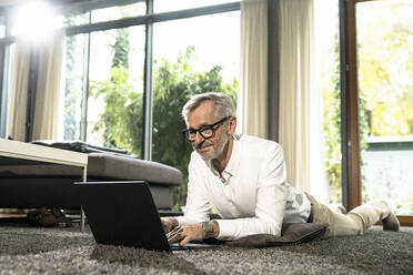 Lächelnder älterer Mann mit grauen Haaren in modernem Design-Wohnzimmer bei der Arbeit am Laptop - SBOF02120